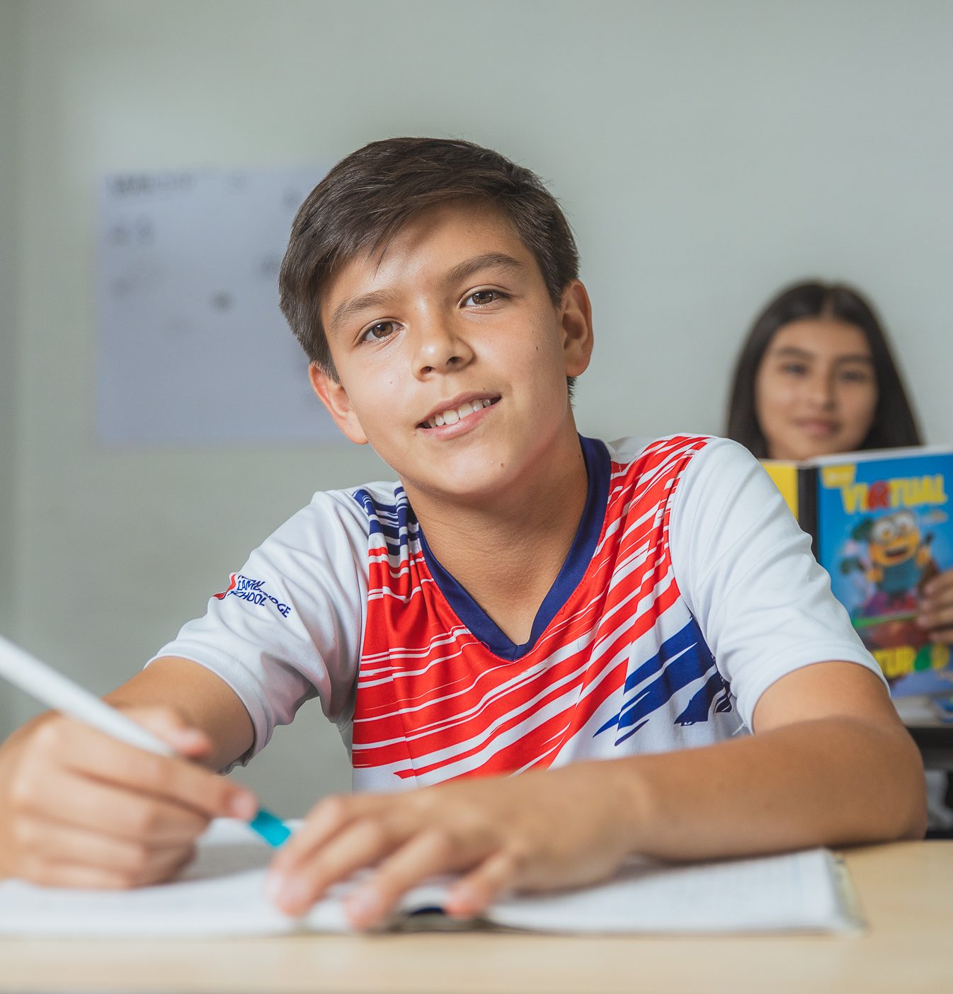 Adolescente mirando a la cámara sonriendo, con un lápiz en la mano derecha y un cuaderno sobre la mesa