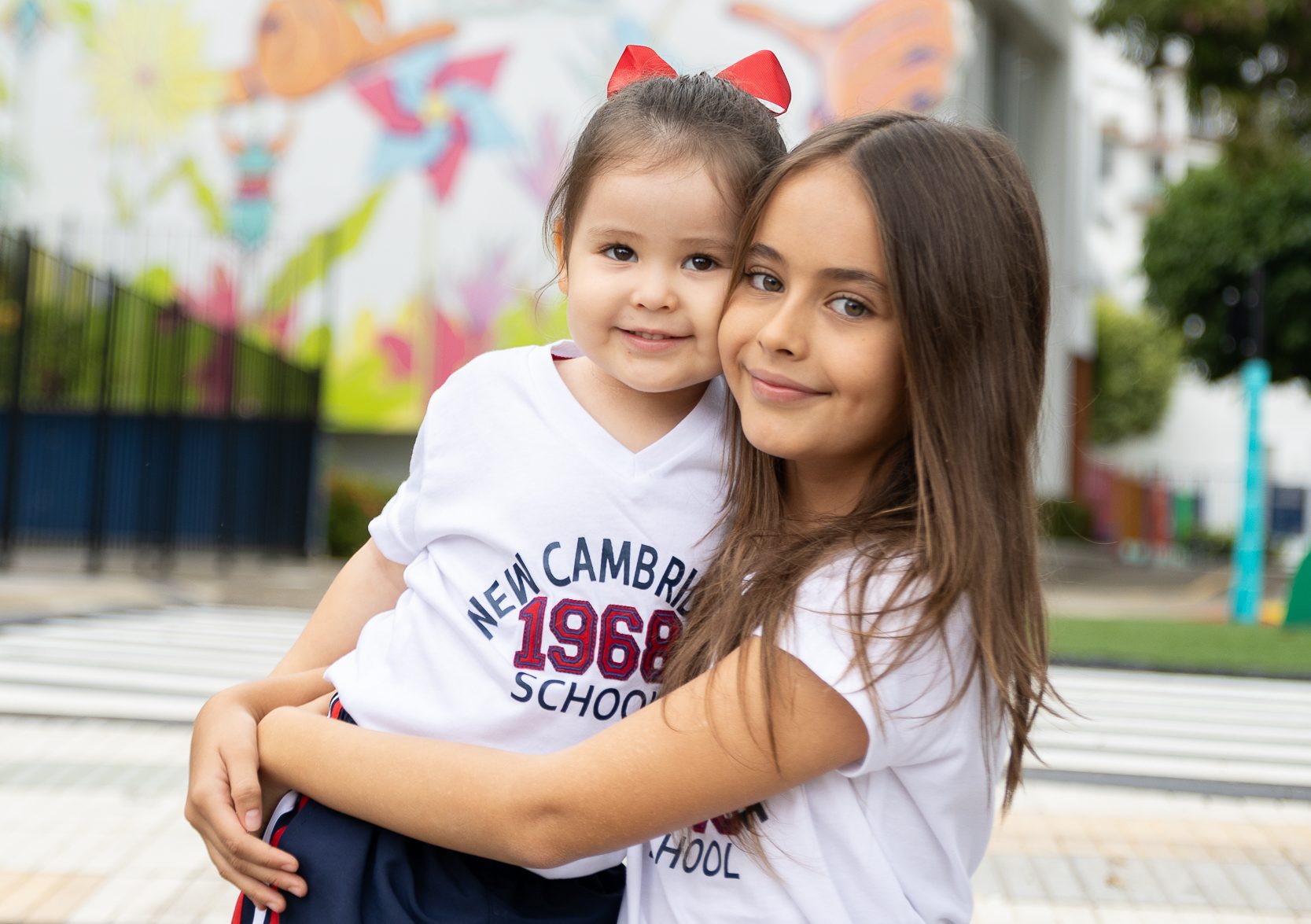 Niña de cabello largo y liso, abraza a una niña mucho menor que ella, de cabello recogido con un moño rojo, ambas tienen camiseta blanca