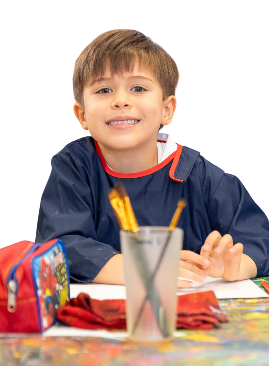 Estudiante de Lower Primary con delantal de artes, en plano medio y con pinceles y cartuchera sobre la mesa