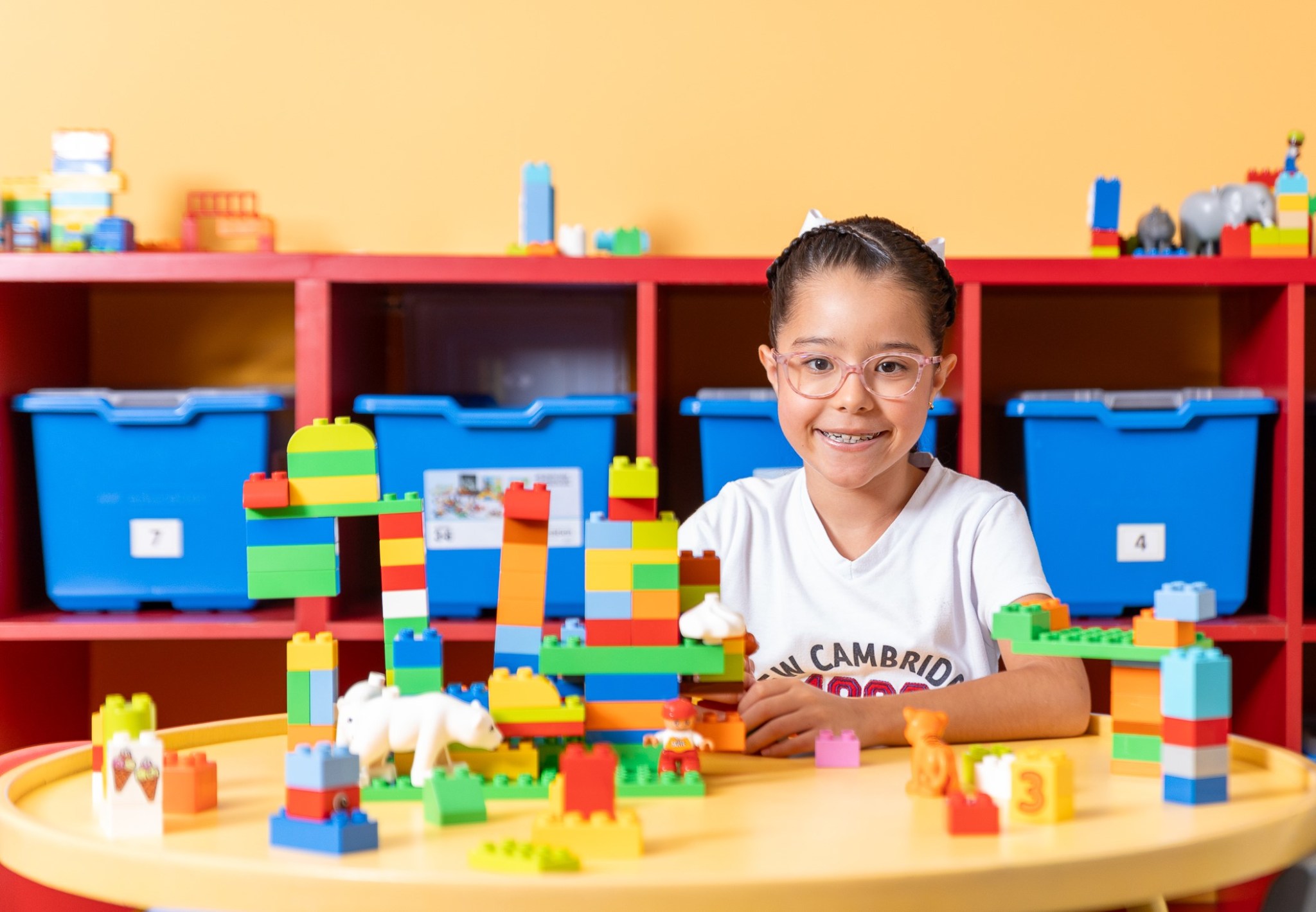 Niña sentada mirando a la cámara y sonriendo, junto a una mesa llena de figuras de lego