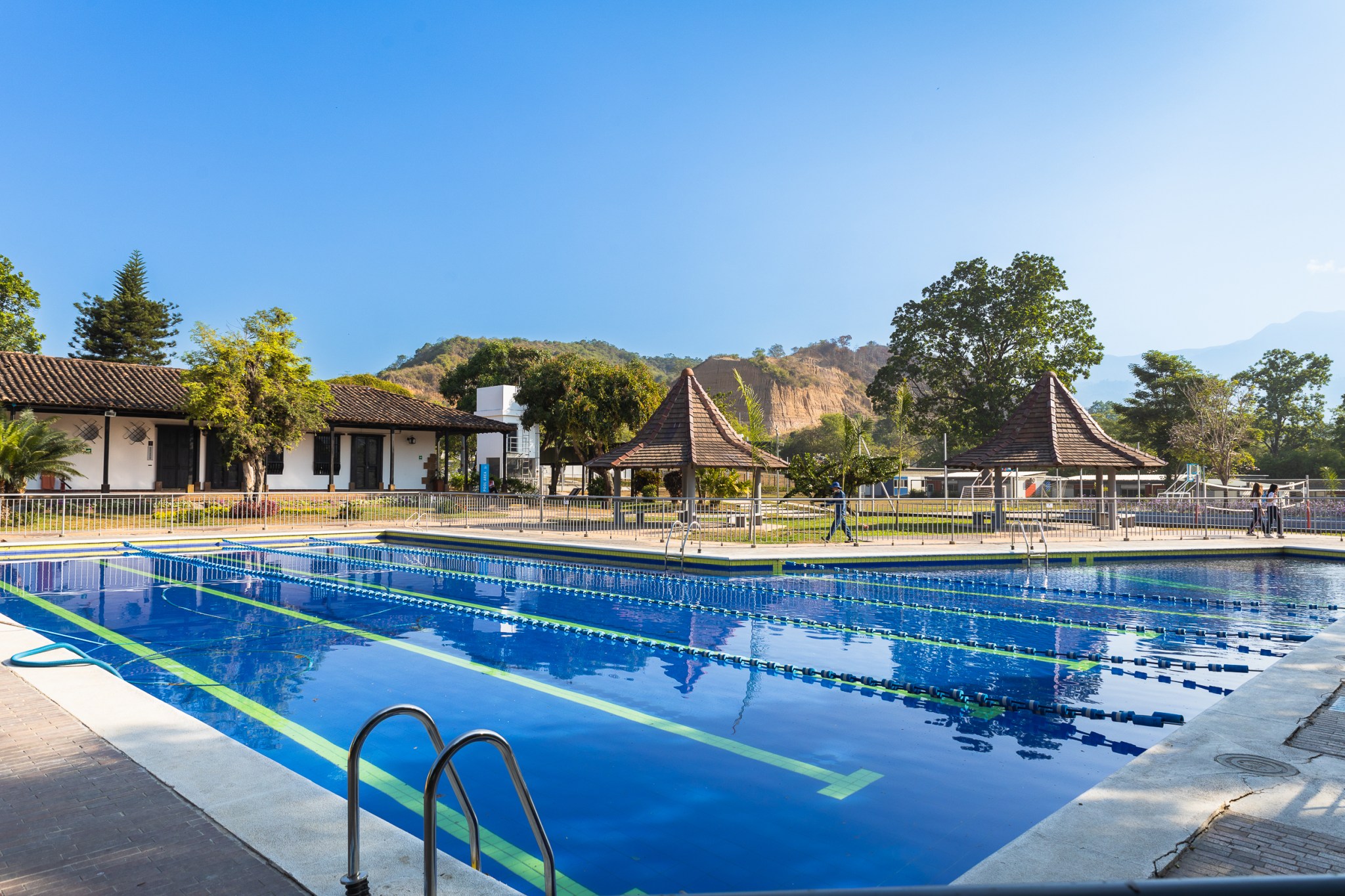 Piscina que se extiende de izquierda a derecha y al fondo se muestran árboles, quioscos y una casa