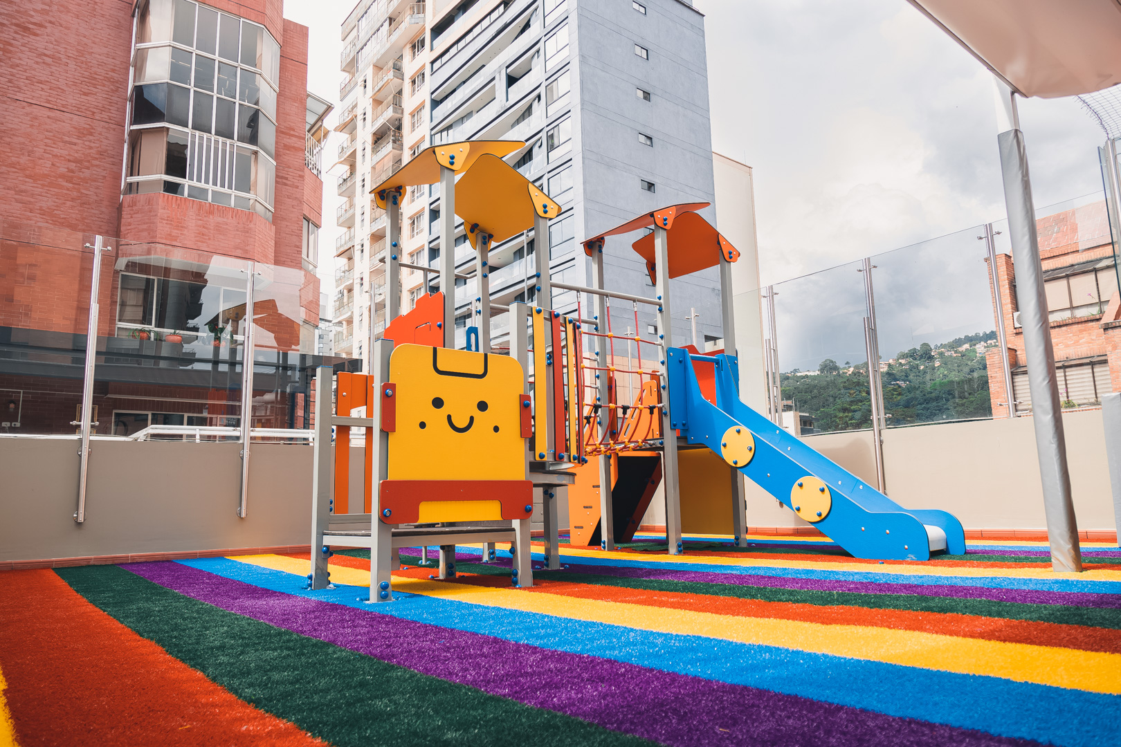 Juego infantil que se encuentra sobre una alfombra de varios colores y al fondo se muestran edificios