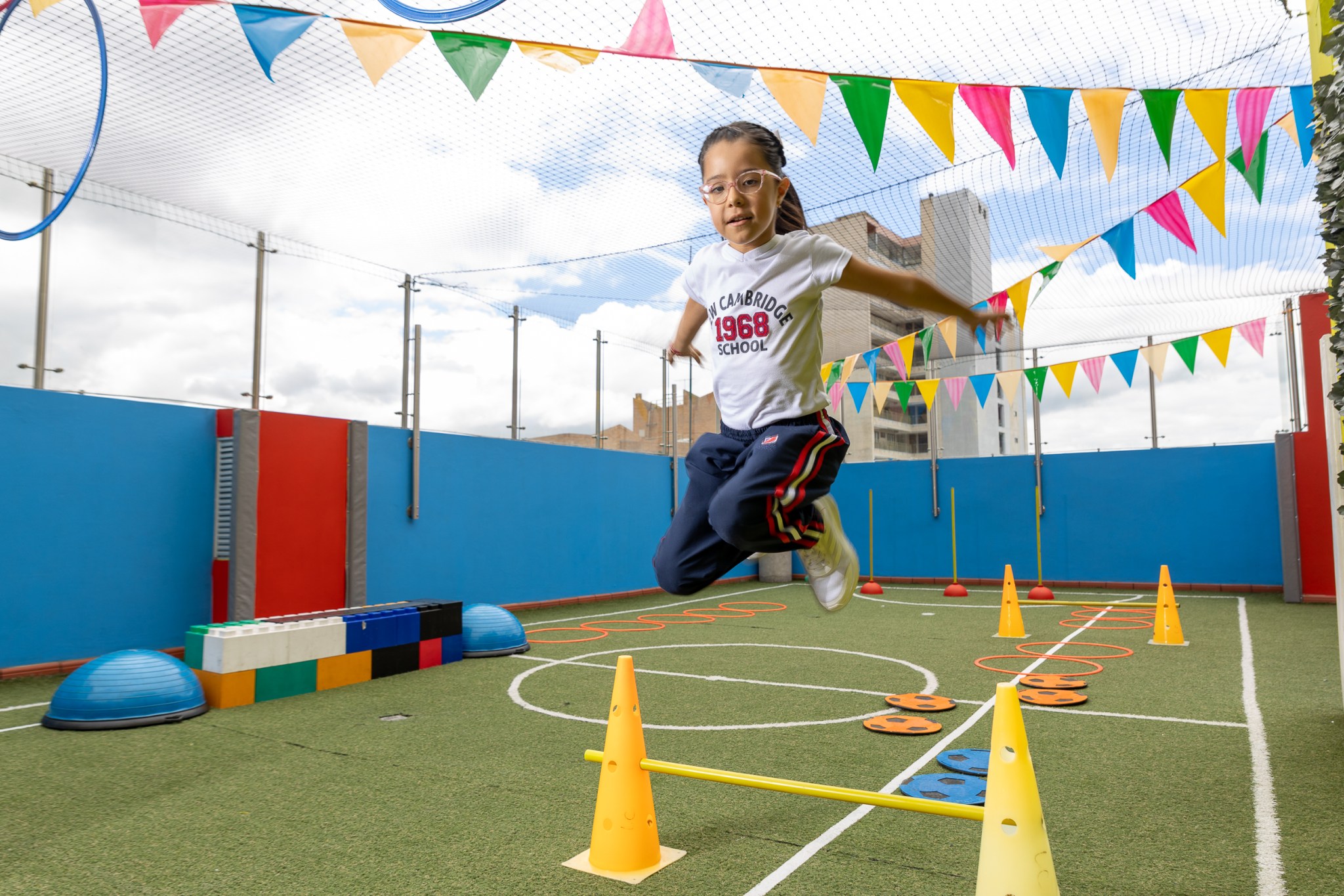 Estudiante saltando en una cancha, en la que hay conos de colores y algunos instrumentos deportivos de colores