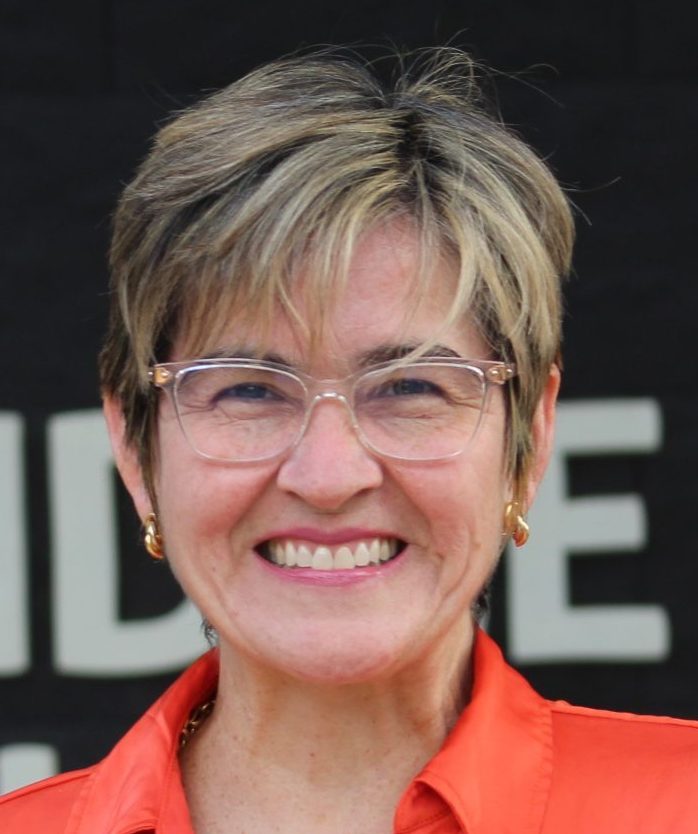 Mujer de cabello corto, rubio y con gafas, con camisa de cuello naranja y cadena en el cuello