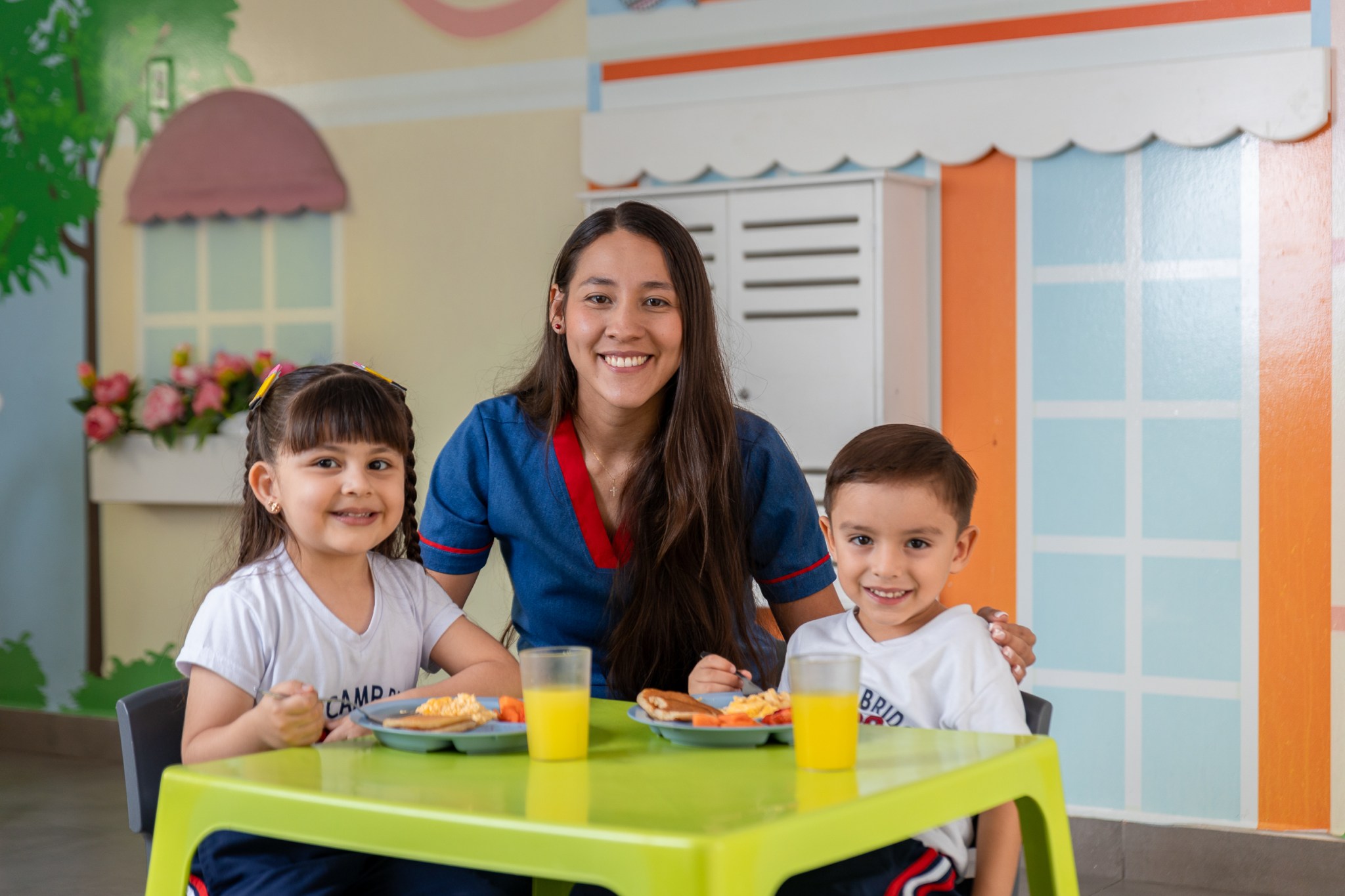 Dos estudiantes en el restaurante de Cabecera site, sentados en una mesa verde, disfrutan de su lonchera y entre ellos los acompaña una docente